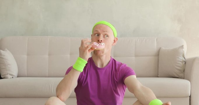 Funny man practicing meditation in lotus yoga pose and eating donoughts at home with funny face. Sportive humor. Camera moving away from face, portrait. Joke, comical, mem, parody, fun behavior.