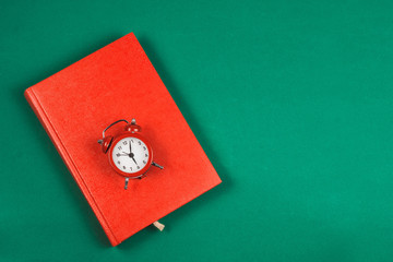 Top view of a red Notepad and a small alarm clock, on a green background. Concept of time and planning. Time to fill out your gratitude diary in the morning. Copy space