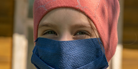 schoolgirl in knitted hat and blue disposable mask looks straight standing against blurry garden close view