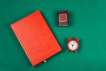 Top view of a red Notepad, a small book and a small alarm clock, on a green background. Romance. Concept of time and planning. Time to fill out your gratitude diary in the morning. Copy space