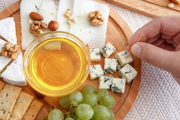 Fototapeta na wymiar Chef`shand holds a piece of cheese. Cheese mix on a wooden plate decorated with grapes, nuts, salad and olives with cheese knifes 