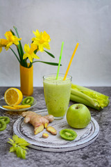 Still life of green smoothie, celery, fruit, ginger root and daffodils in a yellow vase on a gray marble background.