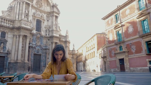 Model sitting on a terrace drinking coffee and looking at the bill with the Murcia Cathedral in the background.