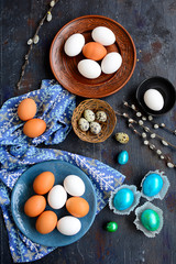 Top view on a dark blue table with unpainted eggs on blue and brown plates and bright blue and green Easter eggs.