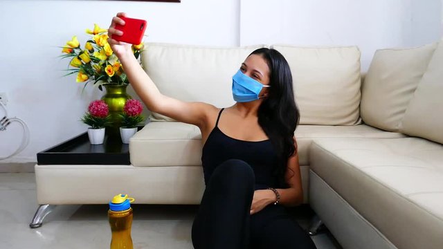 Portrait of a young brunette woman with face mask using her mobile phone while sitting at home after a physical workout
