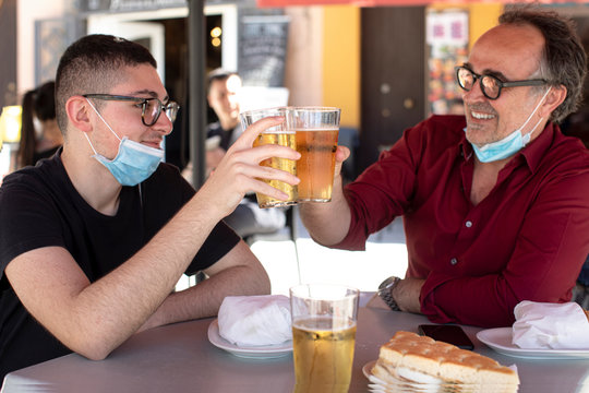 padre e figlio con mascherina brindano al tavolo con un boccale di birra felici per la ritrovata libert&agrave;