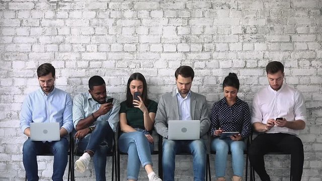 Diverse young people sitting on chairs in row line queue with gadgets, waiting for interesting vacancy job interview. Happy mixed race seekers applicants preparing for meeting with hr manager.