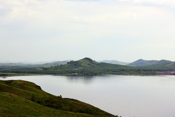 Fototapeta premium Panoramic view. A lagoon with a clean mountain lake in the midst of majestic mountains in a haze of fog. Green grass. Apartments around the lake