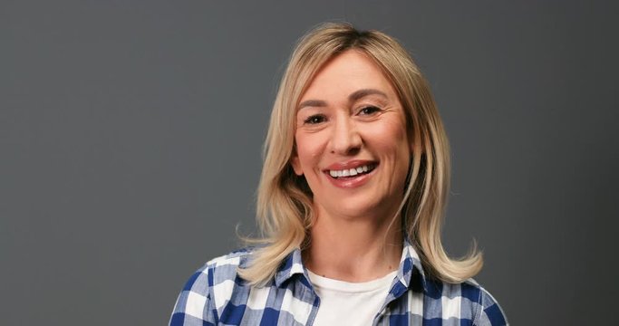 Beautiful cheerful blonde female in good mood standing on grey background in studio. Close up of joyful emotional Caucasian middle-aged woman smiling to camera indoors. Portrait concept