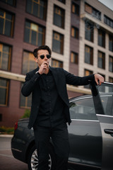 A stylish businessman smokes cigars near a luxury car. Fashion and business