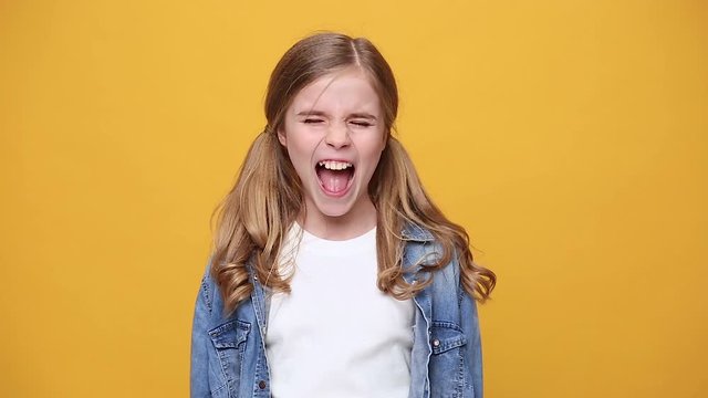 Little angry blonde kid teen teenager girl 12-13 years old in denim jacket white t-shirt isolated on yellow background studio. People childhood lifestyle concept Looking camera scream shout scolding
