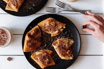 Woman's hand decorates pancakes with chocolate, kiwi and banana. Black plate, white table. From above