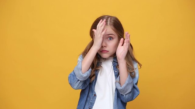 Scared shocked fun little kid girl 12-13 years old in denim jacket white t-shirt isolated on yellow background studio. Childhood lifestyle concept Looking at camera afraid cover hiding face with hands