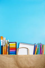 Fototapeta premium Multi-colored pastels, pencils, brushes and a white notebook in a paper bag on a blue background. hobby and art store concept. flat lay, copy space, vertical