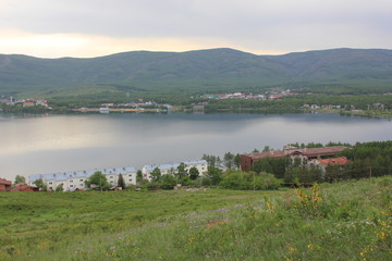 Obraz premium Great panoramic view. A lagoon with a clean mountain lake in the midst of majestic mountains in a haze of fog. Green grass. Apartments around the lake