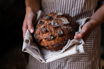 Concept of homemade bread, small bakery, natural farm products, domestic production. Healthy, tasty organic food. Woman in apron holding round whole grain bread freshly baked in her hands. Close up