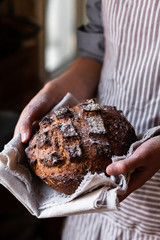 Concept of homemade bread, small bakery, natural farm products, domestic production. Healthy, tasty organic food. Woman in apron holding round whole grain bread freshly baked in her hands. Close up