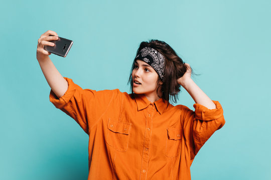 Confident beautiful young woman taking selfie and posing. Hold one hand behind another. Smartphone in another hand. Isolated over blue background.