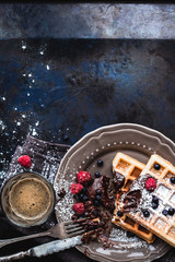 Belgian waffles with chocolate, berries and powdered sugar, a hand with a knife and fork, glass cup with coffee in the process of eating. Brussels breakfast.