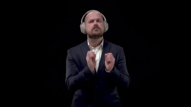 Bald bearded handsome businessman, man in blue jacket and white shirt in headphones, rejoices in victory, and merrily and energetically dances on black background.