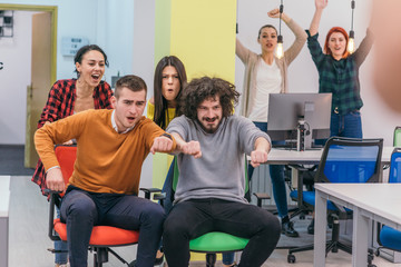 Team building and office fun.Four young cheerful businesspeople in smart casual wear having fun while racing on office chairs and smiling..