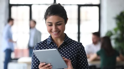 Head shot friendly young indian female manager using digital computer tablet in office. Smart biracial businesswoman working on electronic device, communicating with clients online at workplace.