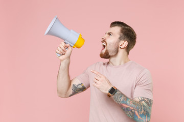 Angry young bearded tattooed man guy in pastel casual t-shirt posing isolated on pink background studio. People lifestyle concept. Mock up copy space. Scream in megaphone, point index finger aside up.