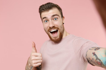 Close up of excited young bearded tattooed guy in pastel casual t-shirt isolated on pink background. People lifestyle concept. Mock up copy space. Doing selfie shot on mobile phone, showing thumb up.