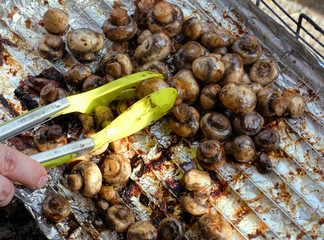 Barbecue sausages and mushrooms grilled on a fire grill and rotating by green kitchen tongs. 