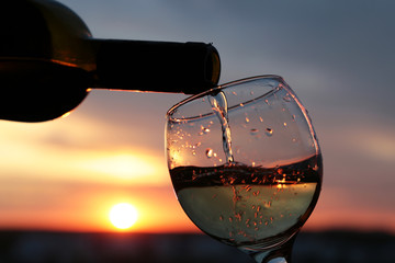 White wine pouring from a bottle into the glass on beautiful sunset background, orange sun is shining through the clouds. Concept of celebration, romantic dinner