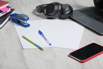 table with paper and stapler on  background of laptop with large black headphones. Notebooks and pencil with pen and mobile phone