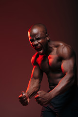 Fototapeta na wymiar Sports shot with a red light of a dark-skinned muscular bodybuilder showing naked torso muscles and screaming