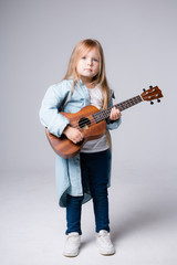 Fototapeta premium A girl a child of British appearance with long hair stands in a blue shirt with ukuleles in his hands on a gray background