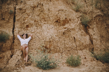 Boho girl in white shirt lying on sandy cliff on beach. Creative aesthetic image. Carefree young woman relaxing on sand seashore. Natural concept. Connection to nature