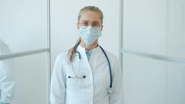 Slow motion of female doctor young blonde standing in white suit and facial mask against white background and looking at camera. People and healthcare concept.