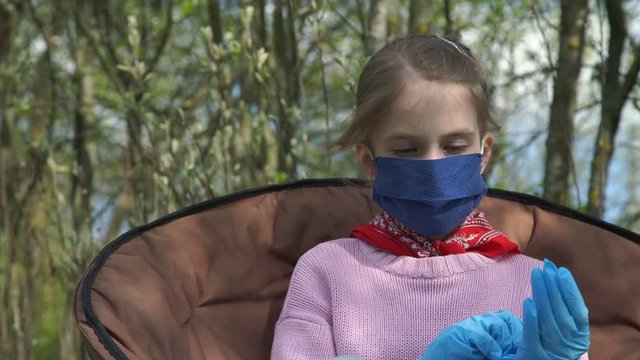 schoolgirl in sterile mask and oversize blue gloves sits in folding armchair being bored against garden slow motion close view