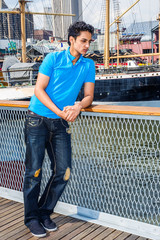 Lonely East Indian American male teenager thinking outside in New York City, wearing blue T shirt, black broken fashionable jeans, cloth shoes, standing by fence at harbor by river, looking down, sad