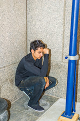 Lonely East Indian American teenager thinking outside in New York City, wearing black long sleeve shirt, broken fashionable jeans, cloth shoes, squatting on corner by wall, fist touching head, sad..