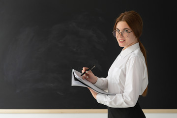 young teacher is standing near a clean blackboard. Keep a blank notebook in your hands.