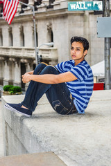 Lonely East Indian American teenager thinking outside in New York City, wearing blue striped T shirt, broken fashionable jeans, cloth shoes, sitting by Wall Street sign, arms embracing legs, sad..