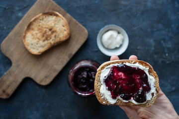 Close up of woman hand holding a sourdough bread toast spread with cream cheese and raspberry jam on blue background
