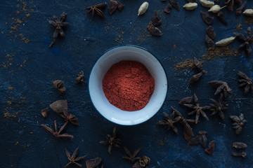 Red chili powder in a small pot surrounded by cloves cardamom, anise on a blue background