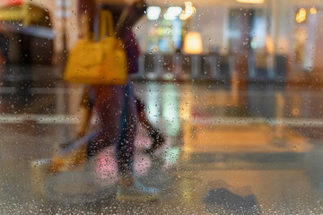 Blurred silhouette of an abstract unrecognizable girl with a handbag, view through a wet window with raindrops, city street in rainy evening