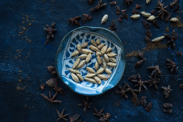 Cardamom pods in a Japanese plate surrounded by other spices like cloves and anise on a blue background