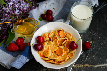 Tiny pancake cereal, cherry, strawberry, honey and milk on dark rustic background with lilac flowers