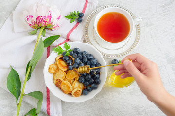 Tiny pancake cereal with blueberry, honey and tea for breakfast in the morning light and pink peony on table