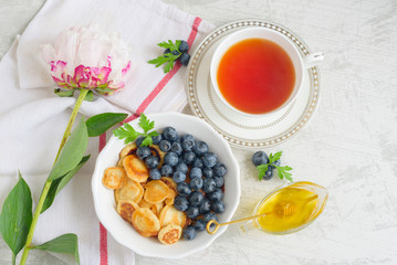 Tiny pancake cereal with blueberry, honey and tea for breakfast in the morning light and pink peony on table