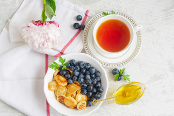 Tiny pancake cereal with blueberry, honey and tea for breakfast in the morning light and pink peony on table