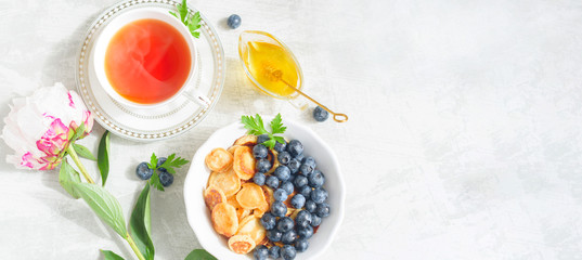 Tiny pancake cereal with blueberry, honey and tea for breakfast in the morning light and pink peony on table