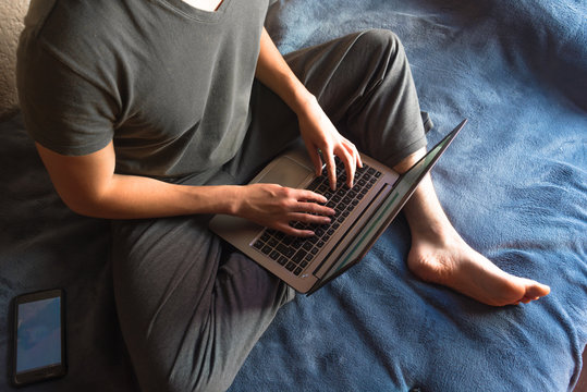 Bird's Eye Angle Photography Of A Homeoffice With A Barefoot Young Man In Gray Pajamas, Studying / Working With A Laptop On His Legs. Dark Blue Blanket Background.